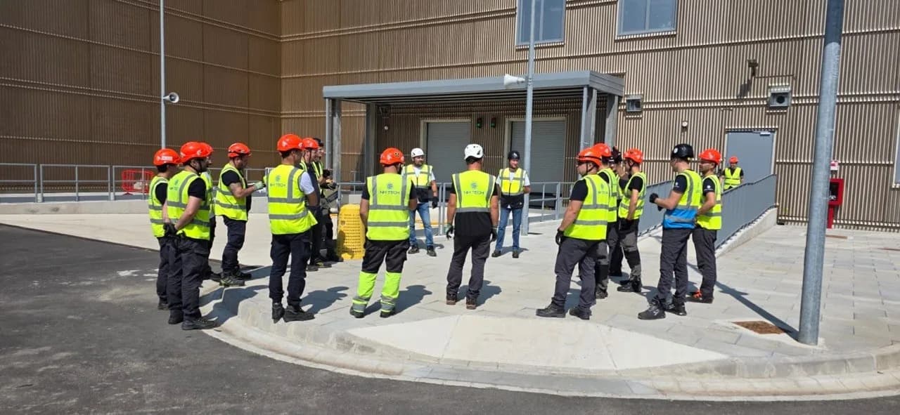 NH TECH technicians in helmets gathered in a circle outside a building entrance with a timber façade