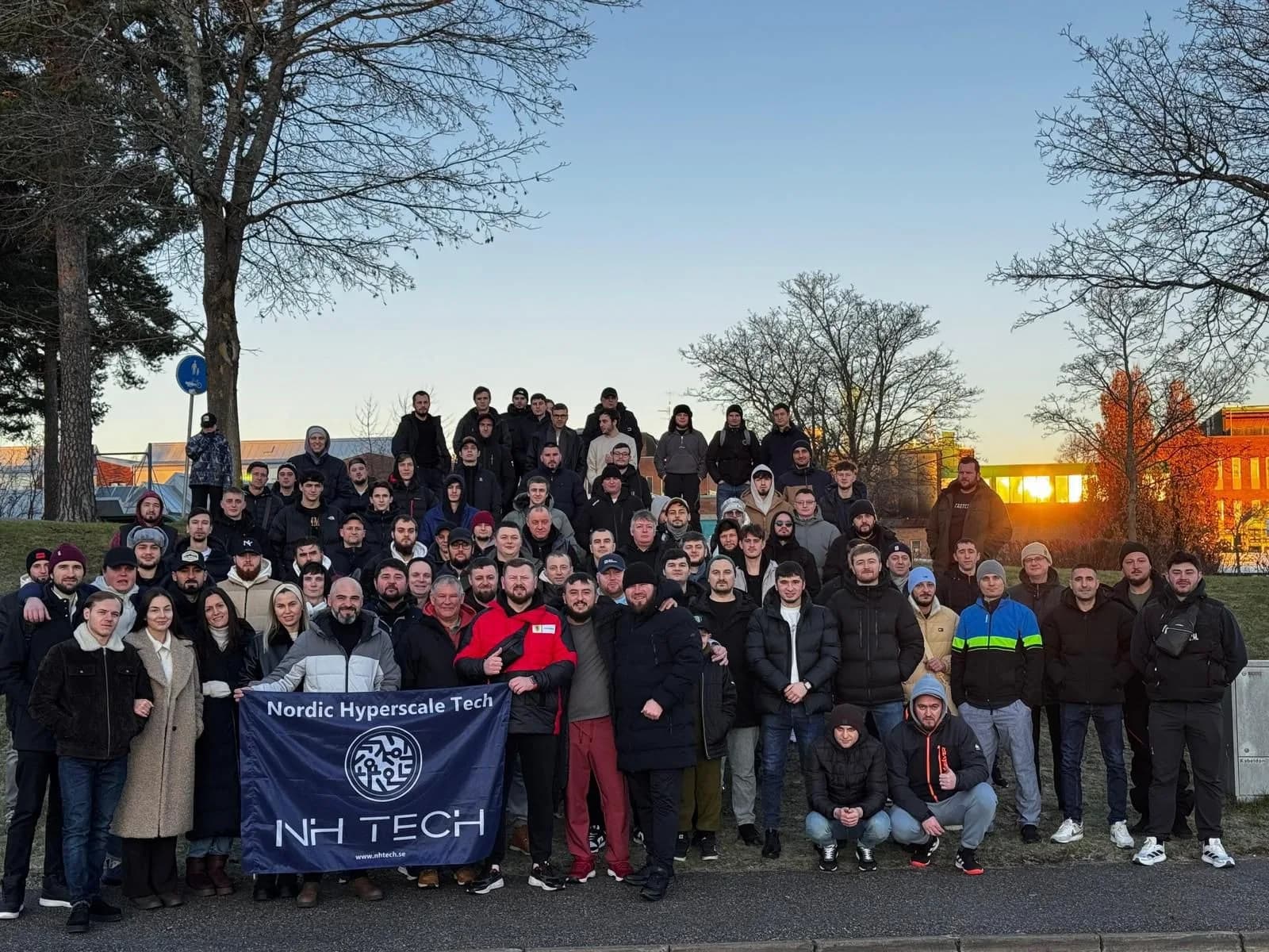 A large group of NH TECH colleagues in winter clothing holding a Nordic Hyperscale Tech banner at sunset