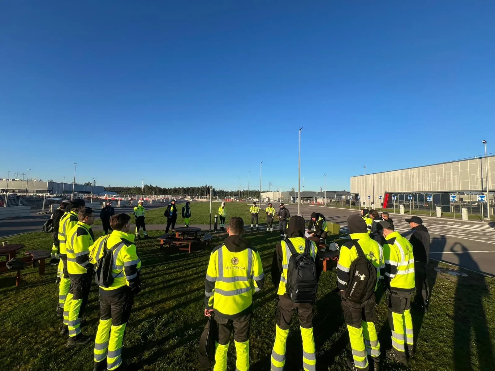 NH TECH technicians in hi-vis jackets standing on grass at sunrise for a morning briefing, data centre buildings behind