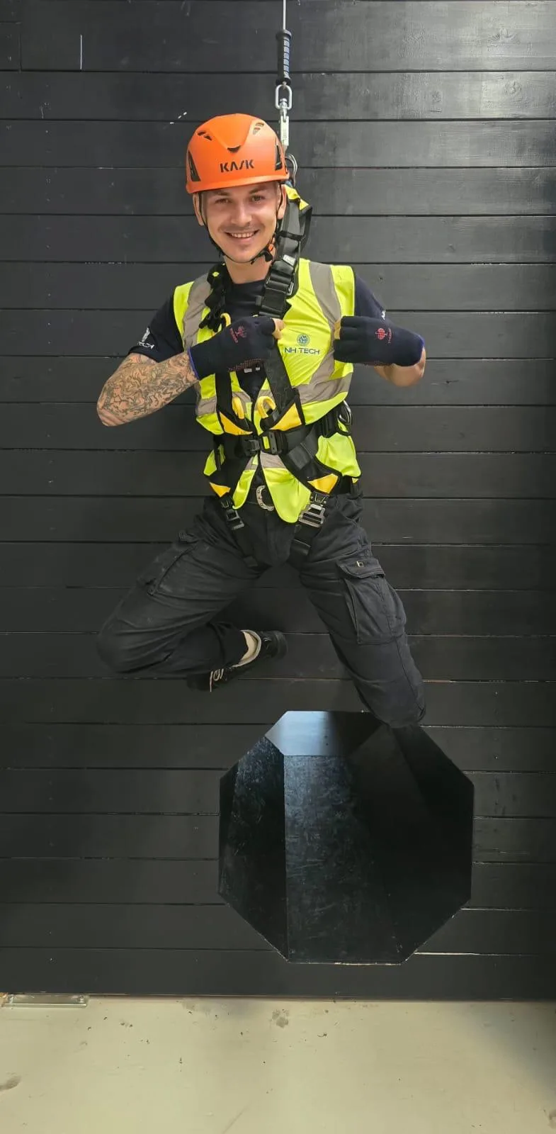 A smiling NH TECH technician in a helmet and full-body harness suspended during working-at-height training