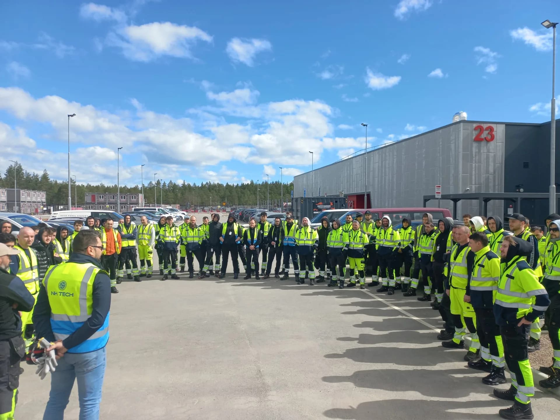 Around fifty technicians in hi-vis standing in a semicircle in a car park listening to a site briefing