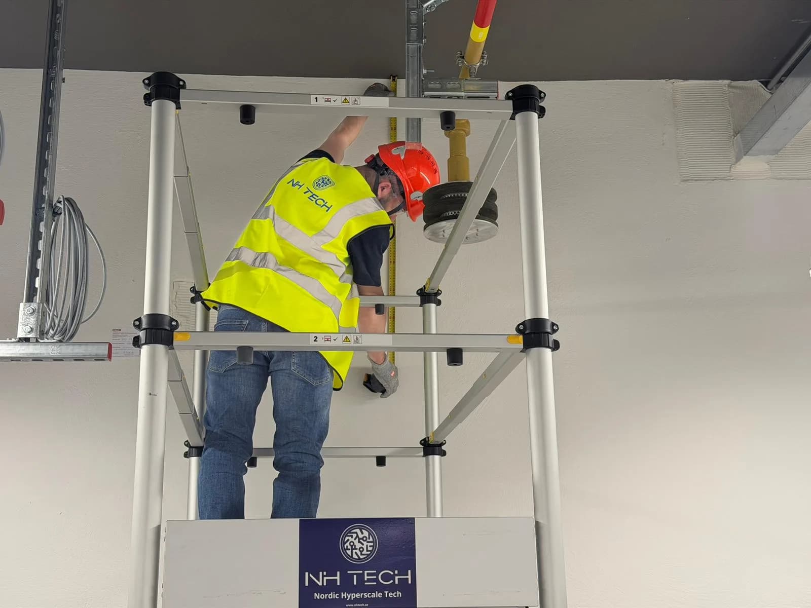 A technician in an NH TECH vest on a podium platform installing a ceiling support, an NH TECH banner on the platform