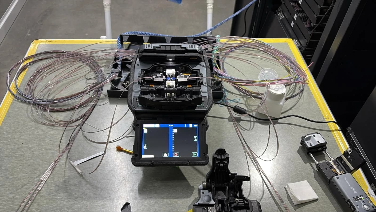 Close-up of a fusion splicer on a work bench surrounded by coiled bare fibres and a cleaver