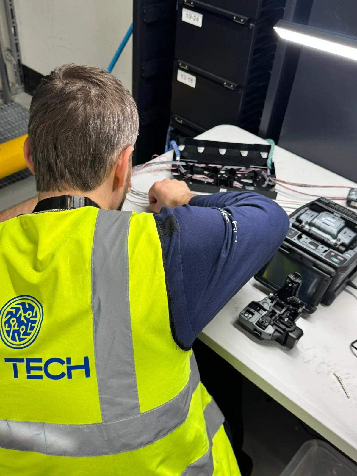 A technician in an NH TECH vest working at a bench with a fusion splicer and an open fibre splice tray